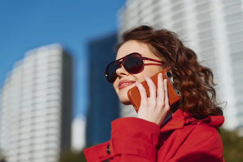 A confident young woman in a red jacket talks on a bright orange phone, wearing sunglasses, with tall modern buildings and blue sky rising behind her as she smiles slightly, suggesting a casual urban
