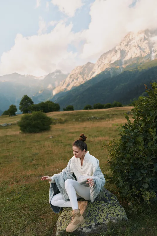 Young woman sits cross legged on a rock in a meadow with distant mountains, wearing a white robe and beige boots, radiating calm and mindful solitude, enjoying quiet solitude.