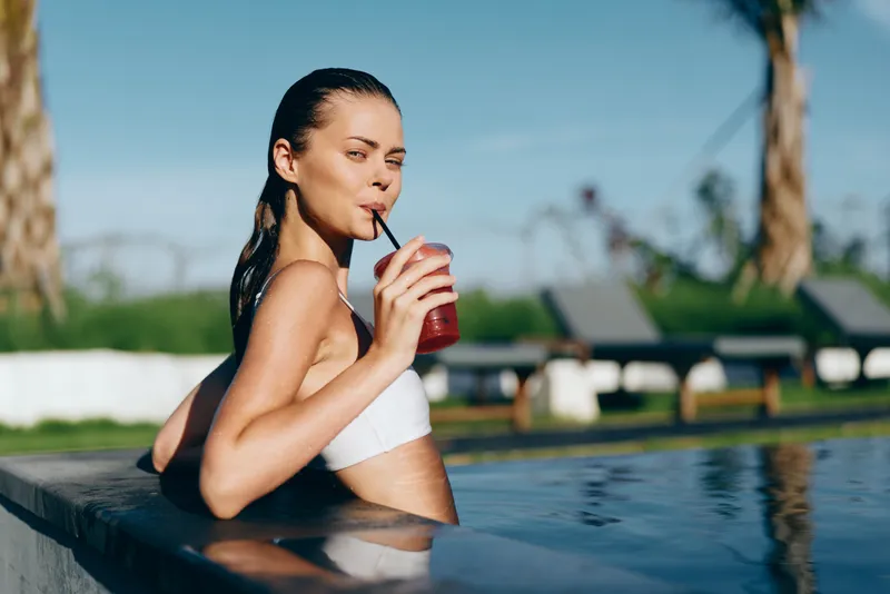 A woman lounges by a poolside, sipping a drink, enjoying a casual summer moment, illustrating outdoor lifestyle and relaxation beside the water.