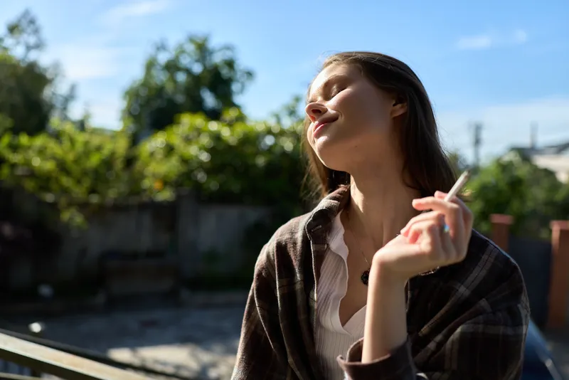 Sunlit woman outdoors with eyes closed and a soft smile, wrapped in a plaid shawl and white blouse, enjoying warm afternoon light and a relaxed, carefree mood of tranquility