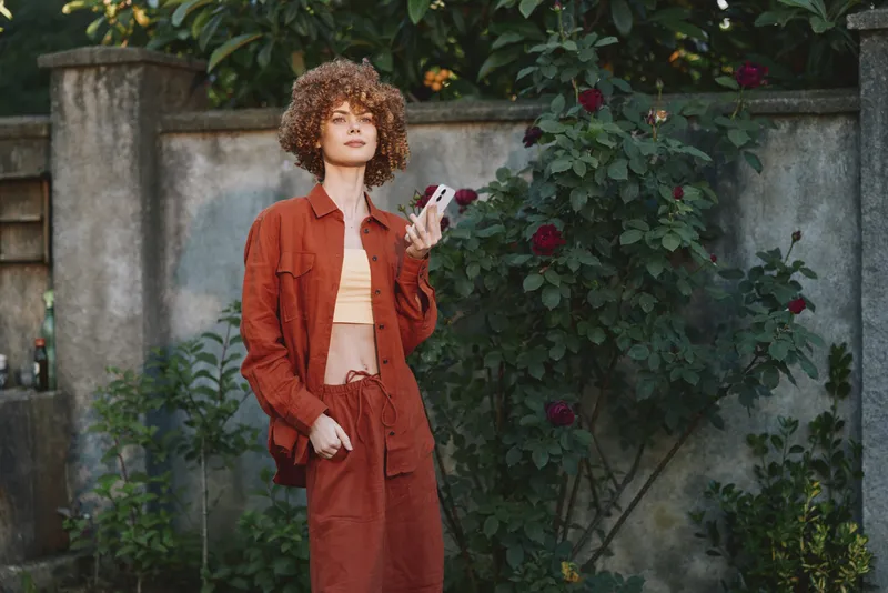 Young woman with curly hair stands in a garden wearing a rust jumpsuit and crop top, looking at her phone with a confident relaxed mood