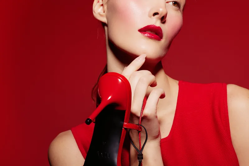 Close-up of elegant woman with red lipstick and red dress holding red high heel shoe near face. Fashion concept with bold makeup and stylish accessories on red background.
