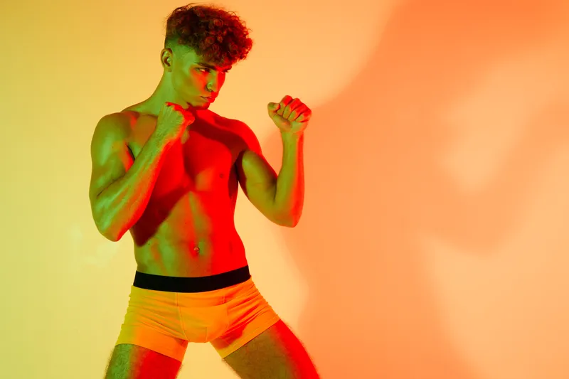 Young muscular man in boxer stance with fists raised, posing shirtless in colorful lighting for fitness, strength, and martial arts training concept in studio environment.