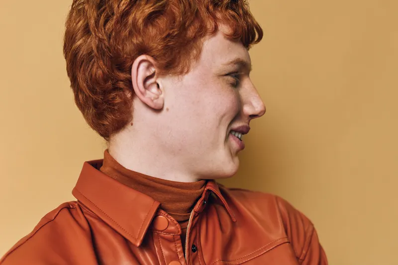 Young man with red hair and freckles is smiling and looking sideways wearing a rust orange shirt and turtleneck against a beige background indoors portrait.