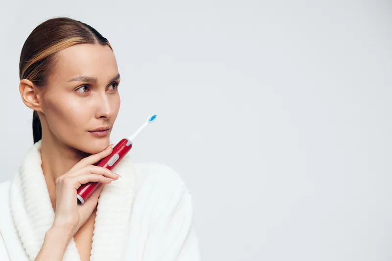 Young woman holding electric toothbrush near face wearing white bathrobe on light background. Concept of oral hygiene, dental care, and morning routine with soft natural lighting.