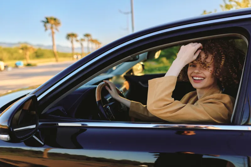 Smiling woman with curly hair driving a sleek car under bright sunlight, palm trees in the background. Casual lifestyle shot highlighting freedom, travel, and everyday moments captured with warm