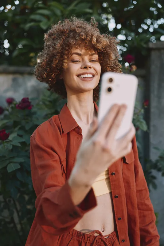 Young woman with curly hair smiles as she takes a selfie outdoors in a casual orange outfit