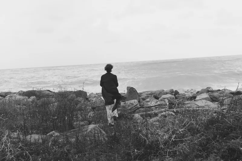 Solitude on a rocky shoreline as a lone figure walks toward the sea, creating a moody seascape with textured rocks, calm water, and a distant horizon in grayscale