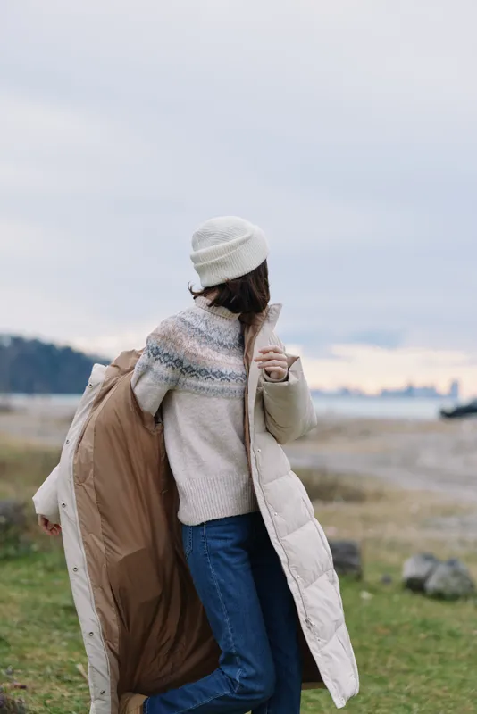Outdoor scene of a person from behind wearing a warm coat and beanie, draping a large coat over the arm, with a calm winter landscape and distant shoreline.