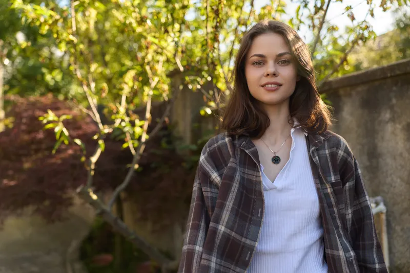 A young woman stands outdoors in a sunlit garden, wearing a plaid shirt over a white top. A relaxed lifestyle portrait conveying casual confidence and fresh, natural style in everyday moments of life