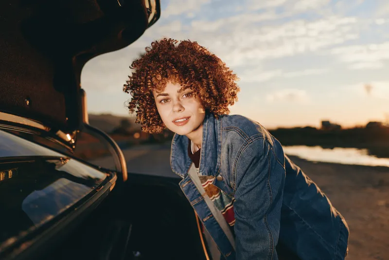 Woman with curly hair is smiling by an open car door at sunset. Outdoors on a road in golden hour, denim jacket, warm backlight and gentle soft wind, casual lifestyle travel mood.