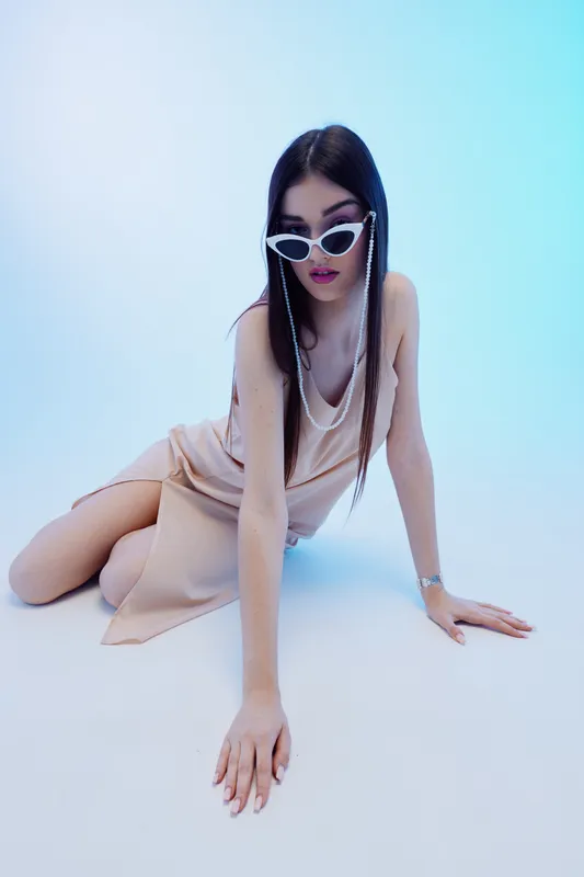 Young woman in beige dress and white sunglasses poses on the floor against blue background. Concept of woman beauty fashion with modern style and elegance.