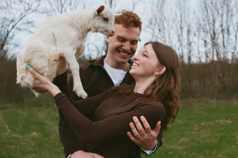 A joyful couple hold a goat kid in a sunny outdoors scene, capturing lifestyle moments with animal companionship, field and nature in a warm, candid portrait of happiness and love.