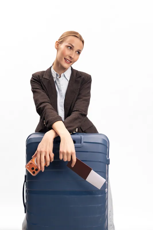 Young woman in business attire leans on large blue suitcase holding passport and phone. White background, travel concept for business trip, vacation, airline, and journey preparation.