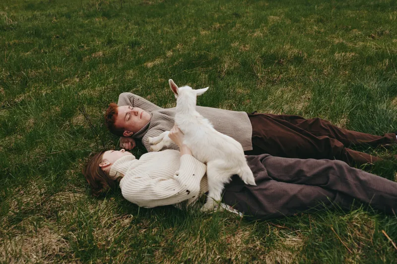 Relaxed couple lying on grass with a curious goat kid climbing nearby. Warm earthy tones highlight outdoor lifestyle, companionship, and gentle play. Soft daylight creates a cozy, candid moment of