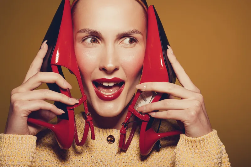 Young woman with red high heels near her face smiles playfully on brown background. Concept of fashion, style, beauty, and joyful emotions in studio portrait.