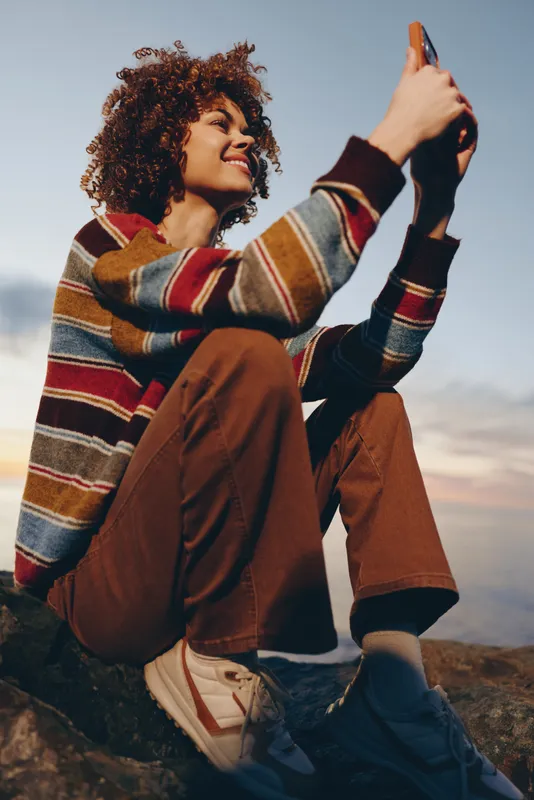 Woman selfie phone outdoors sunset, curly hair in striped sweater and brown jacket, smiling at phone screen while sitting on rocky ground, warm evening light, casual lifestyle