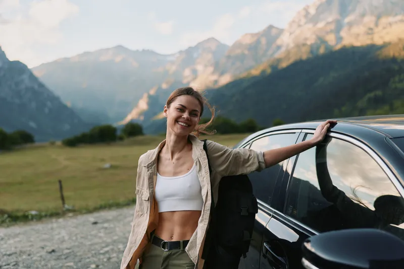 Cheerful woman outdoors by a car, smiling with sunlit mountains in the background, casual outfit, relaxed pose, fresh breeze, natural tones, vibrant travel vibe