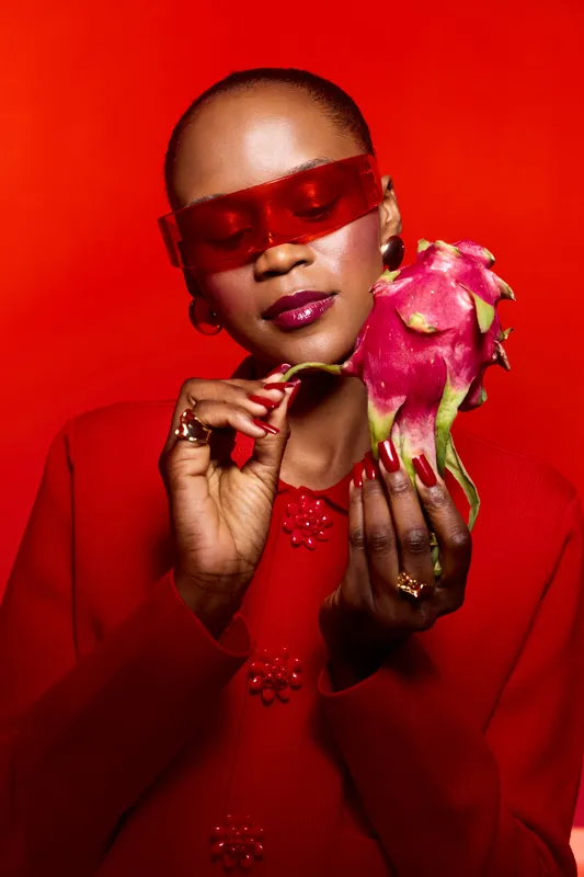 Elegant dark skinned African American woman in red outfit and red sunglasses holds dragon fruit with closed eyes on red background. Concept of fashion, beauty, fruit, and vibrant colors.