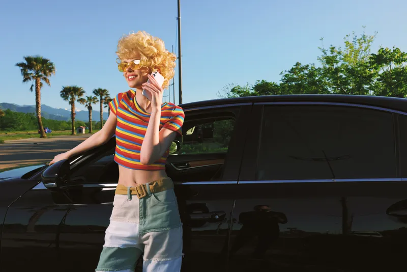 A stylish woman with a curly blonde wig leans on a sleek black car, wearing a striped crop top and denim shorts in bright retro tones. Concept: bold fashion and carefree summer lifestyle.