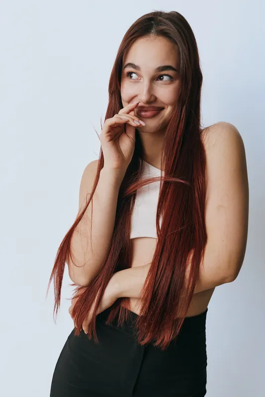 Young woman with long red hair smiling shyly and looking sideways wearing sleeveless white top and black pants on plain white background. Casual style and natural expression.