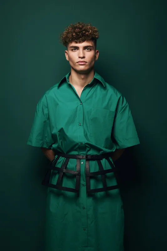 Portrait of young man with curly hair wearing modern bulky black belt over green button shirt posing against dark studio background fashion casual style concept.
