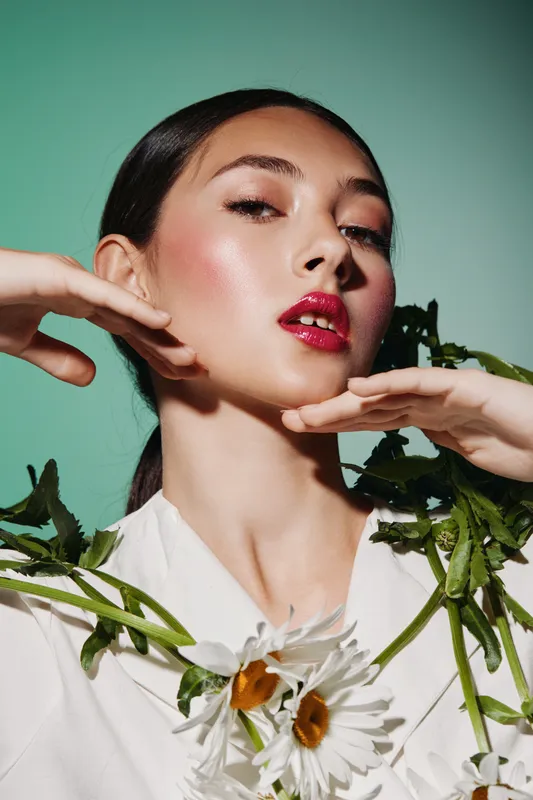 Close-up of fashionable young woman with radiant glowing skin and red lipstick posing creatively with flowers against green background. Concept for beauty fashion editorial.