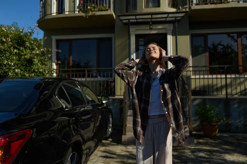 Portrait of a woman enjoying outdoor lifestyle, wearing casual plaid shirt and jeans, standing by a car on a sunny urban street, conveying relaxed mood, confidence and happiness
