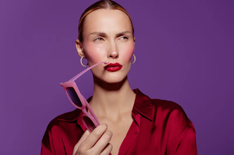 Woman in red blouse holds pink sunglasses near lips, looking away with thoughtful expression. Purple background and hoop earrings complete stylish modern portrait concept of fashion and attitude.