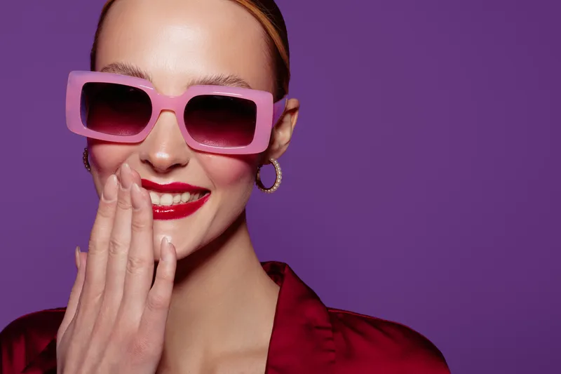 Smiling woman with pink sunglasses and red lipstick covers mouth with hand. She wears red satin blouse and gold hoop earrings on purple background. Concept of fashion and beauty.