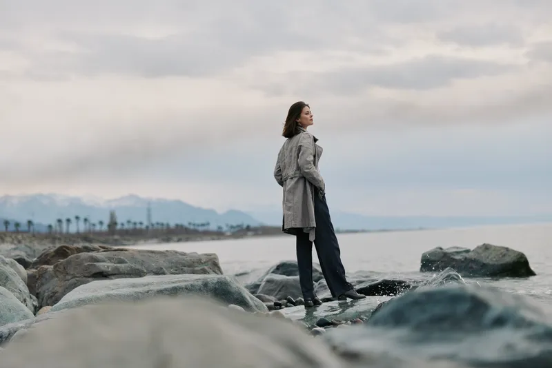 A confident woman stands on rocky shore looking at sea under cloudy sky. The scene shows contemplation and calm with natural landscape in the background.