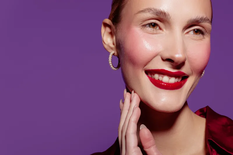 Close-up of smiling woman with red lipstick and hoop earrings touching her face on purple background. Concept of beauty, skincare, makeup, and happiness.