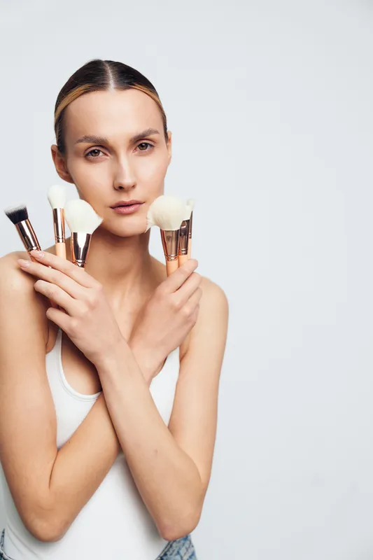 Young woman with natural makeup holds several white makeup brushes with rose gold handles crossed in front of her chest isolated on white background, looking confidently at camera.
