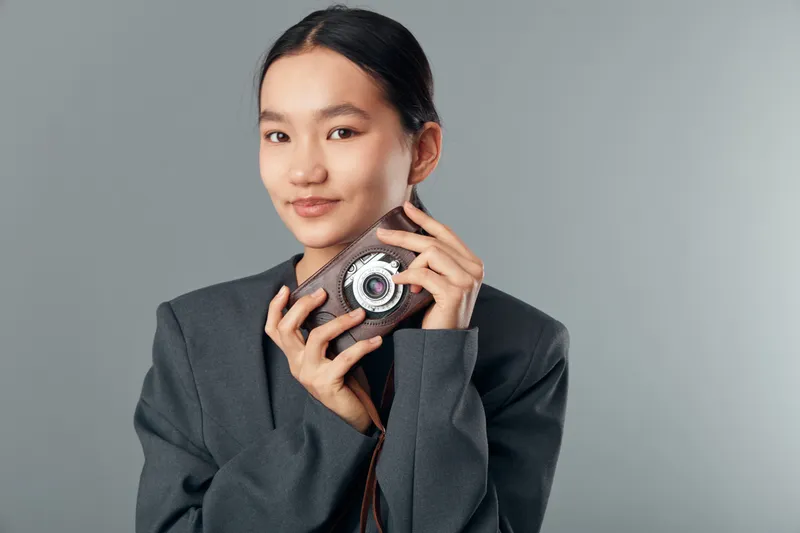 Woman holding vintage camera in hands smiling with confidence, young female portrait in gray jacket on neutral background studio, retro photography and style concept.