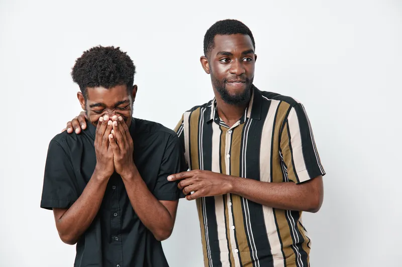 Two African American men with dark skin laughing and joking together in casual clothes on white background. Friends, happiness, and playful moment with smiling young males indoors.