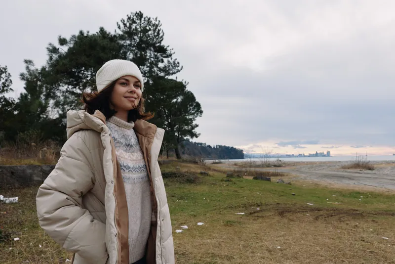 Woman in a warm coat and beanie stands in a windy coastal landscape with trees and distant water, calm expression, casual winter outdoor scene highlighting comfort and nature