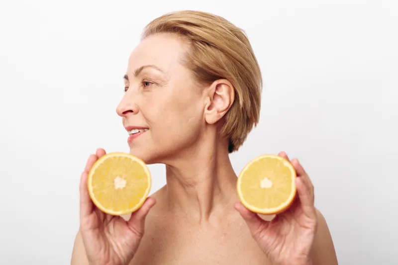 Mature woman with short blonde hair holds two halves of a fresh grapefruit near face, looking sideways. Concept of healthy lifestyle, natural beauty, skincare, and wellness.