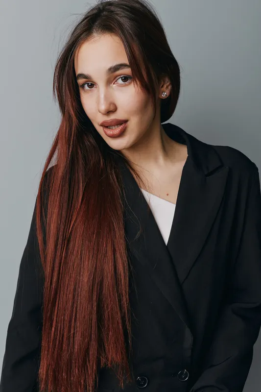 Young woman with long straight hair wearing black blazer and white top posing in studio with neutral gray background. Portrait of confident female model with natural makeup and earrings.