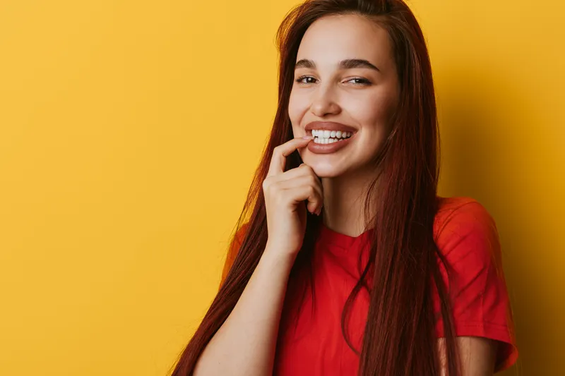Woman smiling with finger near mouth wearing red shirt on yellow background. Portrait of happy young female with long hair and natural makeup in casual style studio shot.