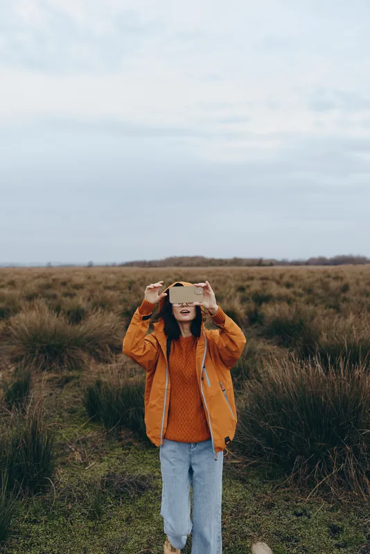 Woman lifestyle outdoors in grassy field, orange jacket and casual jeans, holds smartphone up to take photo, smiling, daylight, soft sky background, wide meadow, portrait, calm mood