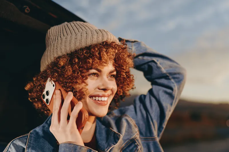 Woman lifestyle outdoors, smiling curly hair, wearing knit hat and denim jacket, holding smartphone to ear, sunny golden hour, gentle breeze, scenic blurred landscape, candid portrait