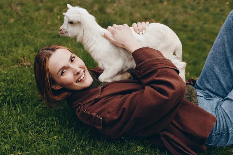 Joyful woman outdoors, lying on grass with a playful goat kid on her chest, warm sunlight, relaxed mood, natural tones, candid moment, gentle companionship, carefree lifestyle