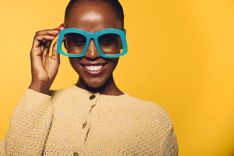 Smiling dark skinned African woman wearing large blue sunglasses and a beige sweater on yellow background. Concept of fashion, style, beauty, confidence, and summer accessories.