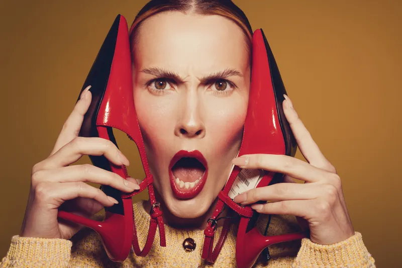 Surprised woman with red lipstick holds red high heels near face on brown background. Concept of fashion, style, emotions, and shopping experience for women.