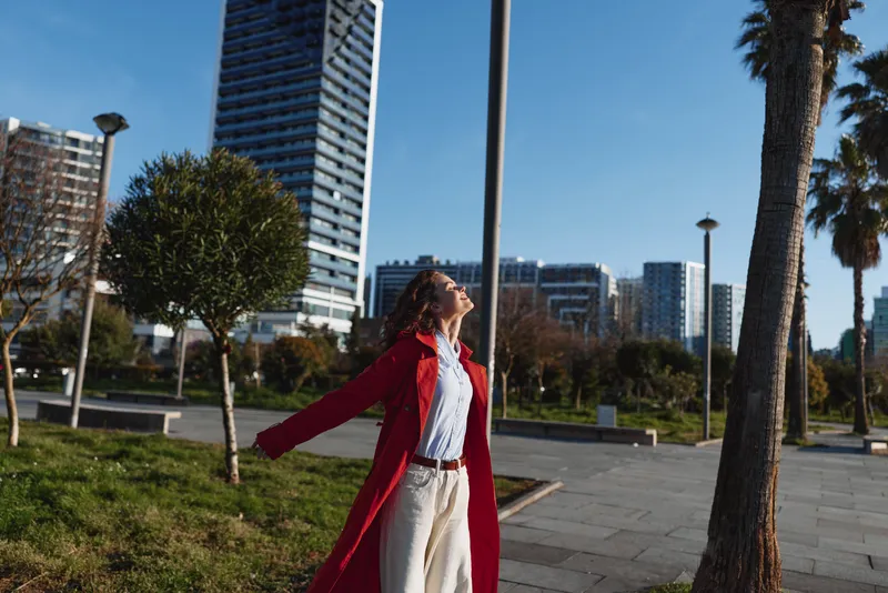 Happy woman in red coat enjoying sunny day outside in urban park with modern buildings in background. Concept of freedom, relaxation, and city lifestyle.