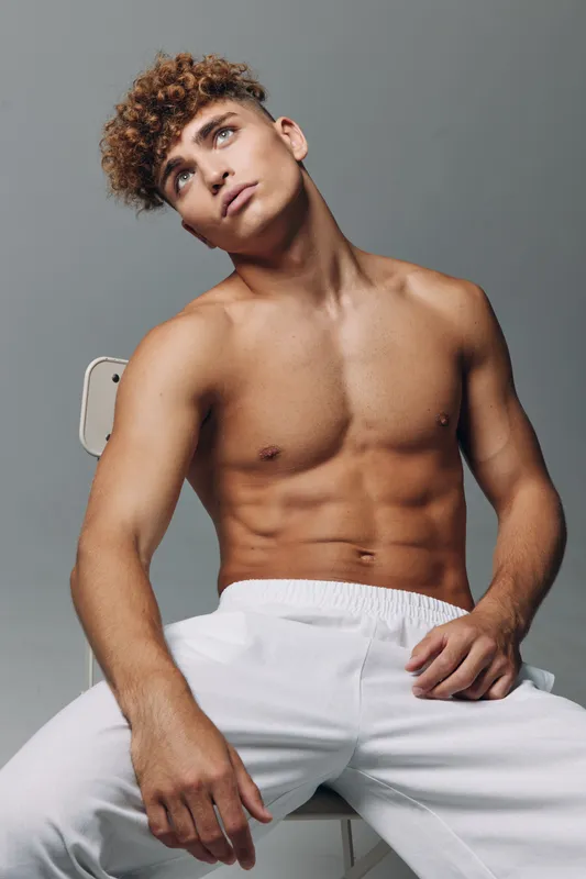 Young shirtless man with curly hair sitting against blank grey background looking slightly upward wearing white pants and exuding casual attitude and relaxed posture in studio.