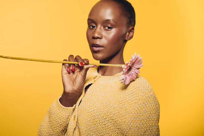 Elegant dark skinned African woman in yellow knitted sweater holds pink flower near face against yellow background. Concept of natural beauty, fashion, and floral aesthetics in studio.