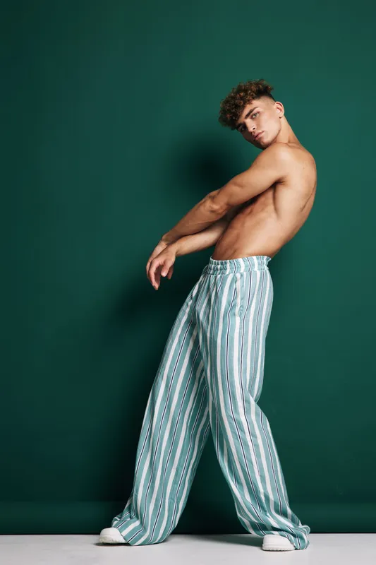 Young man with curly hair posing shirtless in striped wide pants and white sneakers against green background in studio. Fashion, casual style and confident expression.