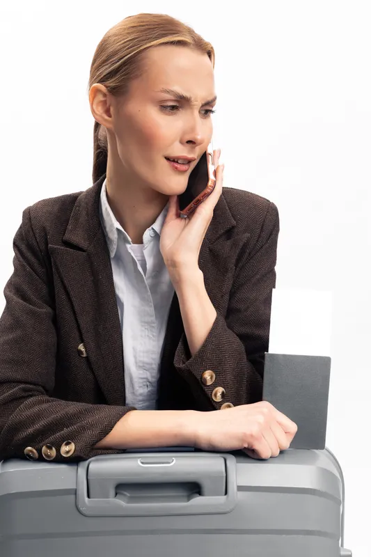 Concerned businesswoman in jacket talks on phone near suitcase with boarding pass. Travel stress and urgent conversation concept with white background and professional attire.
