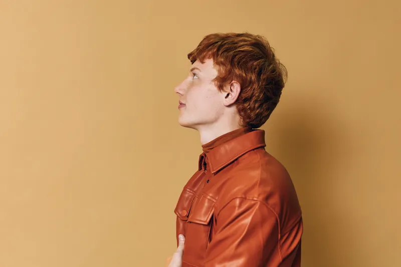 Profile of young man with red hair and freckles in brown leather jacket looking forward on beige background in studio. Side view portrait of thoughtful person with casual style and calm expression.